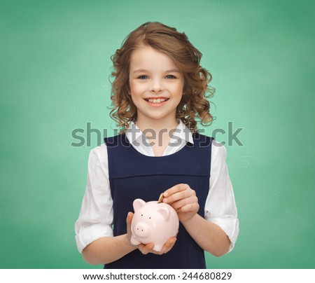 people, money, savings, investments and children concept - smiling school girl putting coin into piggy bank over green chalk board lights background