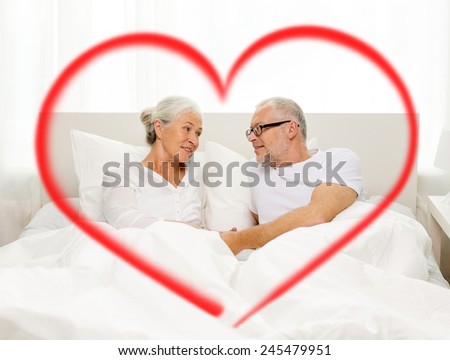 family, bedtime, valentines day, love and people concept - happy senior coupler lying in bad and talking at home with big red heart shape
