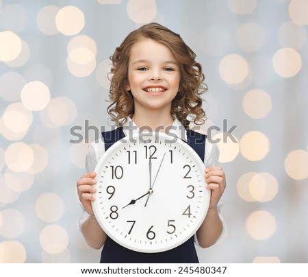 people, time management and children concept - smiling girl holding big clock showing 8 o\'clock over holidays lights background
