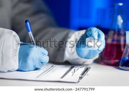 science, chemistry, medicine and people concept - close up of young scientist with chemical sample taking notes on clipboard and making test or research in laboratory