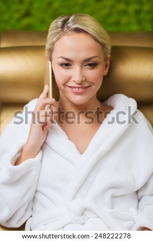 people, beauty, lifestyle, technology and relaxation concept - beautiful young woman in white bath robe with smartphone calling and talking at spa