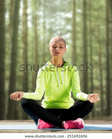 sport, fitness, yoga and people concept - happy young woman meditating in lotus pose and sitting on mat over woods background