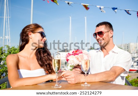 love, dating, happiness and people concept - smiling couple wearing sunglasses with bunch of flowers and champagne glasses looking to each other at cafe