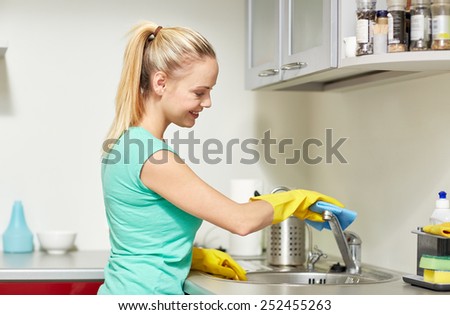 people, housework and housekeeping concept - happy woman in protective gloves cleaning tap with rag at home kitchen