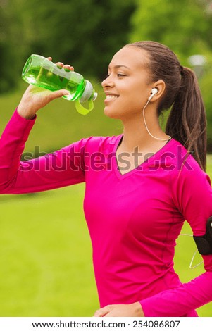 fitness, park, drink and sport concept - smiling african american woman with player and earphones drinking from bottle outdoors