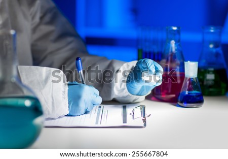 science, chemistry, medicine and people concept - close up of young scientist with chemical sample taking notes on clipboard and making test or research in laboratory