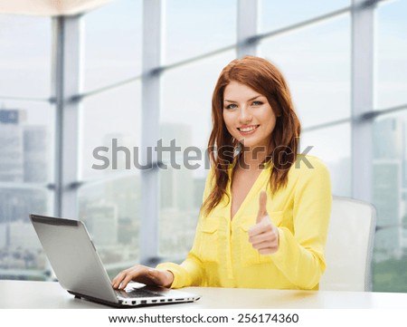 people, business and technology concept - smiling young woman with laptop computer showing thumbs up and sitting at table over office window background