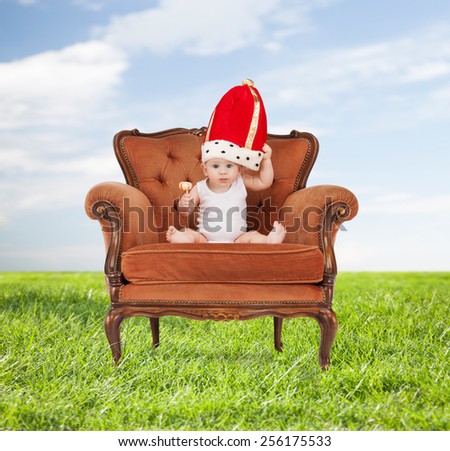 people, childhood and royalty concept - happy baby boy in royal hat with lollipop sitting on chair over blue sky and grass background