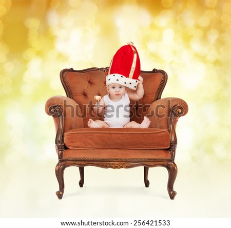 people, childhood, holidays and royalty concept - happy baby boy in royal hat with lollipop sitting on chair over yellow lights background