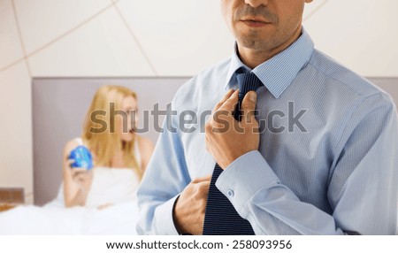 people, business, morning and work concept - close up of man in shirt dressing up and adjusting tie on neck over bedroom with woman holding alarm clock background