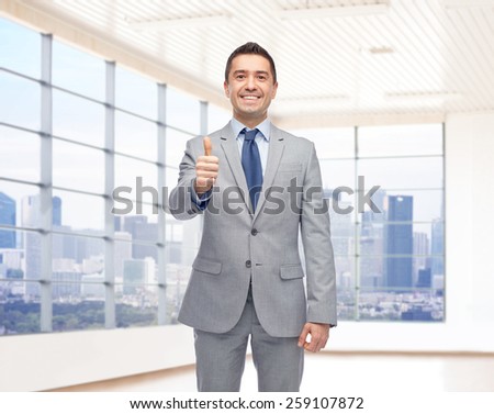 business, people, gesture, real estate and success concept - happy smiling businessman in suit showing thumbs up over city office window background