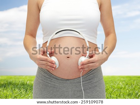 pregnancy, people, music, technology and expectation concept - close up of happy pregnant woman applying headphones to bare tummy over blue sky and grass background
