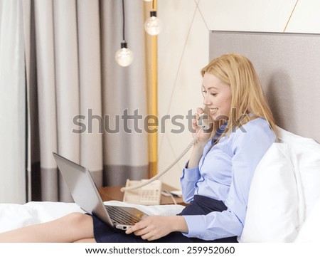 business, technology, internet and hotel concept - happy businesswoman with phone and laptop lying in hotel in bed