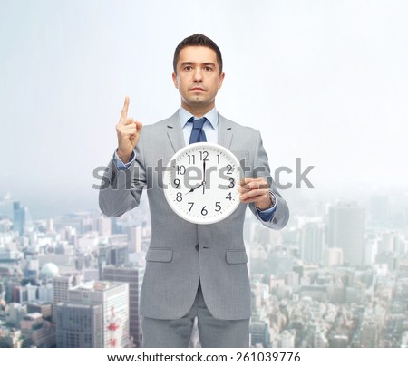business, people, time management and gesture concept - businessman in suit holding clock showing 8 o\'clock and pointing finger up over city background