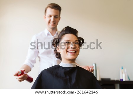 beauty, hairstyle and people concept - happy young woman with hairdresser with hair spray fixating hairdo at salon
