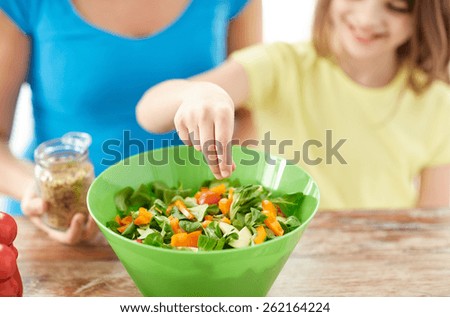 food, healthy eating, family and people concept - close up of happy girl and mother cooking salad for dinner and adding spices in kitchen