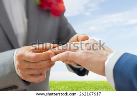people, homosexuality, same-sex marriage and love concept - close up of happy male gay couple hands putting wedding ring on over blue sky and grass background