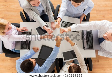 business, people, technology and team work concept - close up of creative team with laptop and tablet pc computers showing thumbs up gesture and sitting at table in office