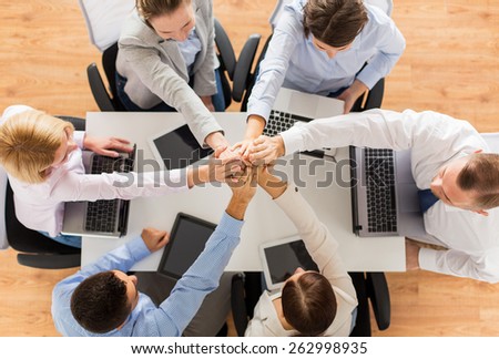 business, people, technology, cooperation and team work concept - close up of creative team with laptop and tablet pc computers sitting at table and holding hands on top of each other in office