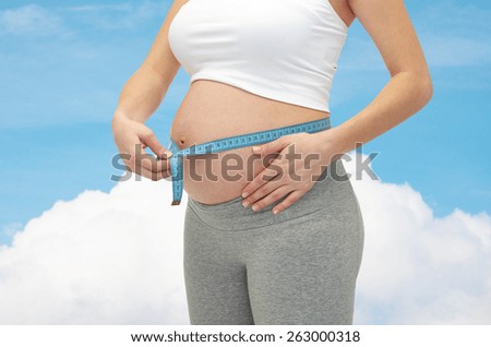pregnancy, motherhood, control, people and expectation concept - close up of happy pregnant woman measuring her bare tummy over blue sky and cloud background