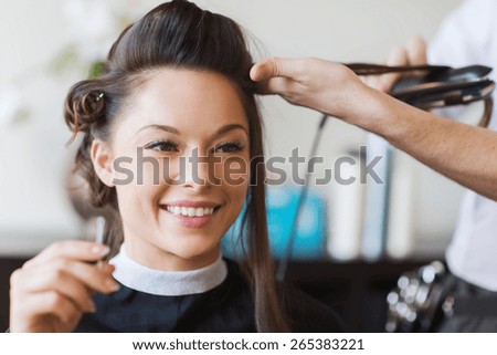beauty, hairstyle and people concept - happy young woman and hairdresser with hair iron making hairdo at hair salon