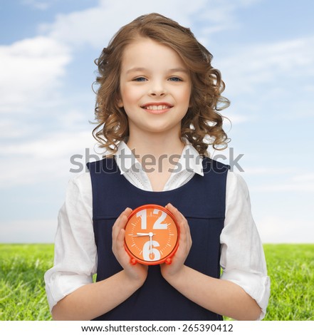 people, childhood, time and punctuality concept - happy girl with alarm clock over blue sky and grass background