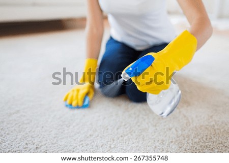 people, housework and housekeeping concept - close up of woman in rubber gloves with cloth and derergent spray cleaning carpet at home