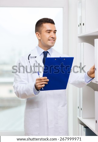 medicine, profession, technology and people concept - happy male doctor with clipboard choosing folder from shelf in medical office