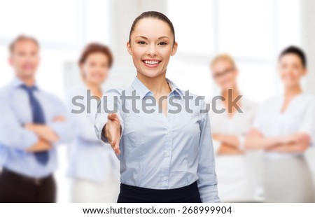 business, gesture and education concept - friendly young smiling businesswoman with opened hand ready for handshake over group of people background