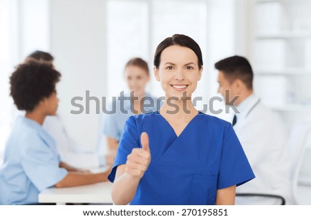 clinic, profession, people and medicine concept - happy female doctor or nurse over group of medics meeting at hospital showing thumbs up gesture