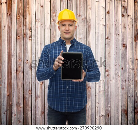 repair, construction, building, people and maintenance concept - smiling male builder or manual worker in helmet showing tablet pc computer blank screen over wooden fence background
