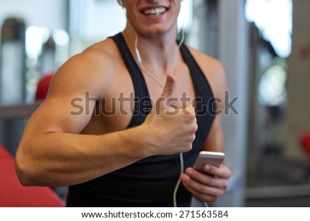 sport, bodybuilding, lifestyle, technology and people concept - happy man with smartphone and earphones listening to music and showing thumbs up in gym