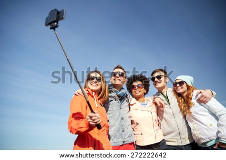 tourism, travel, people, leisure and technology concept - group of smiling teenage friends taking selfie with smartphone and monopod outdoors