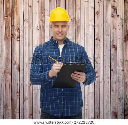repair, construction, building, people and maintenance concept - smiling male builder or manual worker in helmet with clipboard taking notes over wooden fence background