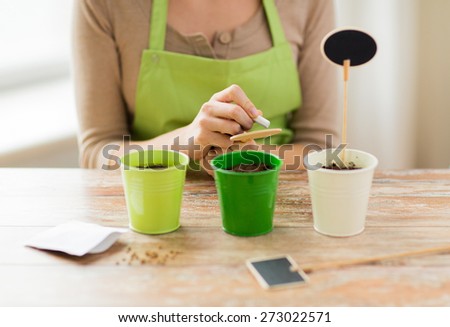 people, gardening, seeding and profession concept - close up of woman writing name on garden sign over pots with soil and seeds