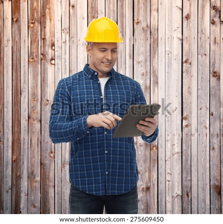repair, construction, building, people and maintenance concept - smiling male builder or manual worker in helmet with tablet pc computer over wooden fence background