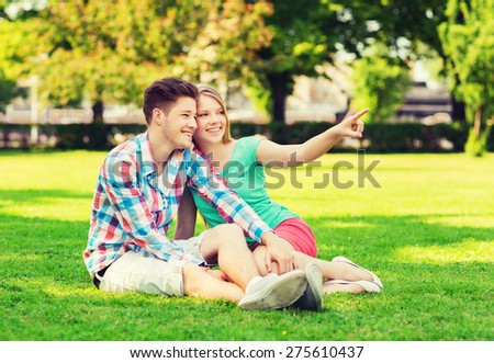 holidays, vacation, love and friendship concept - smiling couple sitting on grass and pointing finger in park