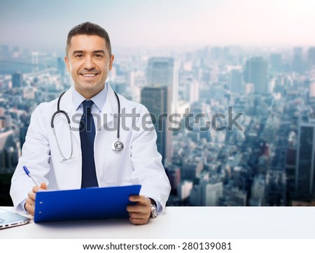 medicine, profession, technology and people concept - happy male doctor with clipboard and stethoscope over city background