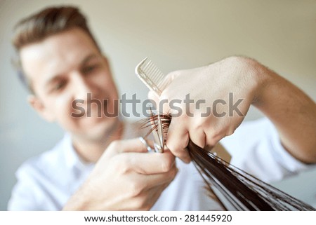 beauty, hairstyle and people concept - male stylist hands with scissors and comb cutting hair tips at salon