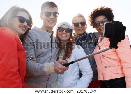 people, leisure, friendship and technology concept - group of smiling teenage friends taking picture with smartphone on selfie stick outdoors