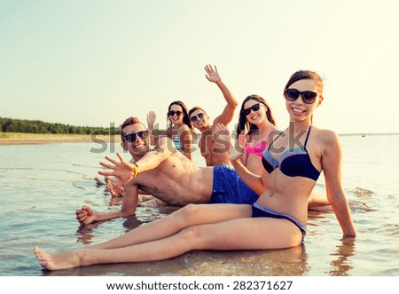 friendship, sea, holidays, gesture and people concept - group of smiling friends wearing swimwear and sunglasses sitting in water on beach
