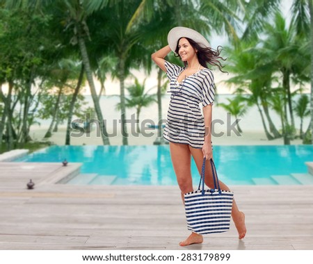 people, fashion, summer and beach concept - happy young woman in summer clothes and sun hat with bag over swimming pool at beach resort