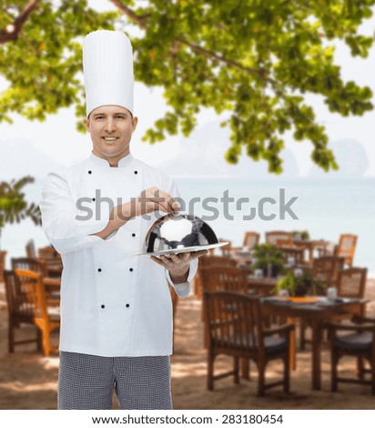 cooking, profession and people concept - happy male chef cook holding cloche over restaurant lounge on beach