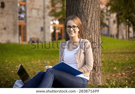 summer, education, learning and teenage concept - happy student girl with laptop computer writing to notebook at campus