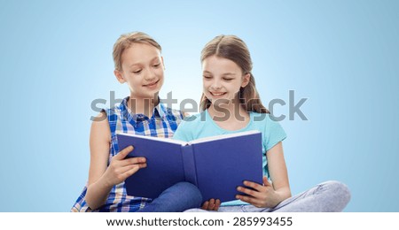 people, children, friends, literature and friendship concept - two happy girls sitting and reading book over blue background