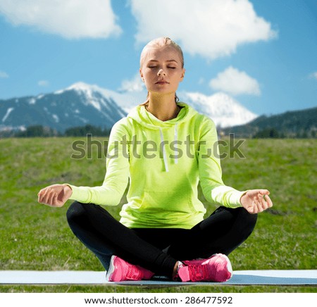 sport, fitness, people and meditation concept - woman sitting in lotus pose doing yoga over mountains, green field and blue sky background