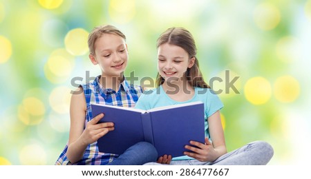 people, children, friends, literature and friendship concept - two happy girls sitting and reading book over green holidays lights background