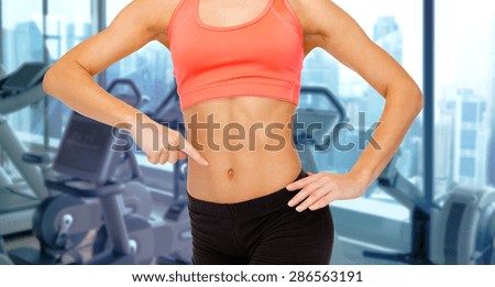 people, fitness and sport concept - close up of woman trainer pointing finger to her abdomen over gym machines background