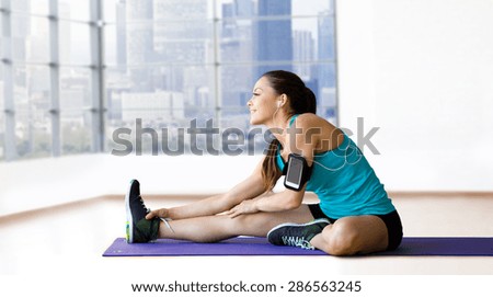fitness, sport, training, technology and people concept - smiling woman with smartphone and earphones listening to music and stretching leg on exercise mat over gym background