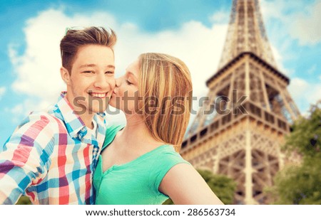 travel, tourism, summer vacation, technology and love concept - happy couple taking selfie with smartphone or camera and kissing over eiffel tower and sky background